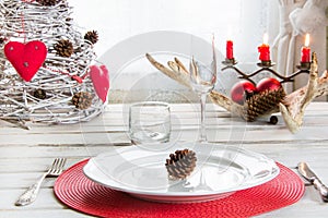 Christmas place setting with white dishware, silverware and red decorations on white board in interior near the window.