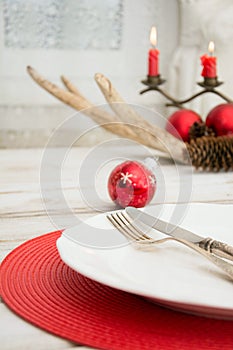 Christmas place setting with white dishware, silverware and red decorations on white board in interior near the window.