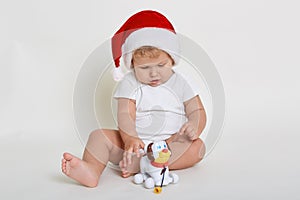 Christmas portrait of charming little baby boy, wearing santa hat, playing with little cute plastic dog toy, infant studying new