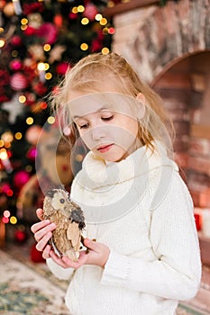 Christmas portrait of happy blonde child girl in white sweater holding toy owl near the Christmas tree and wooden toy horse. New Y