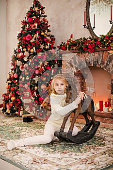 Christmas portrait of happy blonde child girl in white sweater siting on the floor near the Christmas tree and wooden toy horse. N