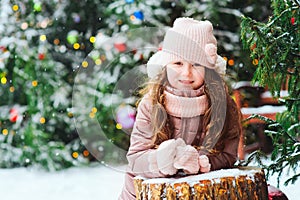 Christmas portrait of happy kid girl playing outdoor in snowy winter day, fir trees decorated for New Year holidays