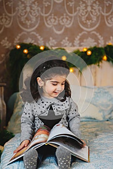 Christmas portrait of happy smiling beautiful little girl sitting on bed, reading book under the christmas tree. Winter holiday