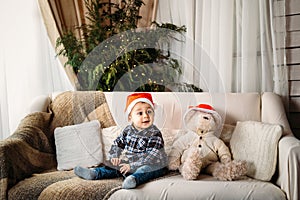 Christmas portrait of happy smiling little boy in red santa hat sitting on sofa playing with toy gun present.