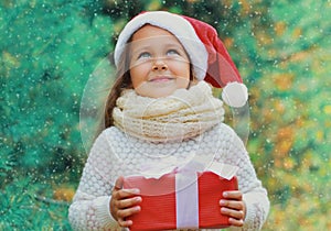 Christmas portrait happy smiling little girl child in santa red hat with gift box near a green branch tree