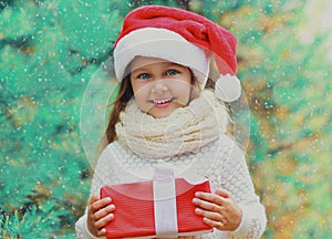 Christmas portrait happy smiling little girl child in santa red hat with gift box near a green branch tree