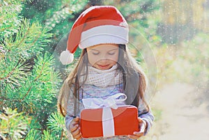 Christmas portrait of happy smiling little girl child in santa red hat with gift box near green branch tree