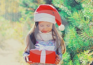 Christmas portrait happy smiling little girl child in santa red hat with gift box near a green branch tree