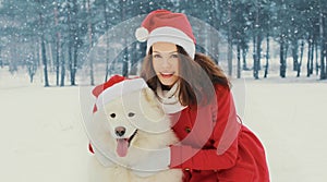 Christmas portrait of happy woman with white Samoyed dog in red santa hat sitting on snow in winter