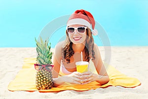 Christmas portrait pretty young smiling woman in red santa hat with pineapple cup fresh fruit juice lying on beach over blue sea