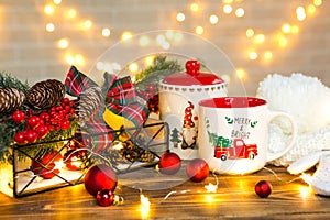 A Christmas red mug with the inscription Merry and Bright and a cookie jar on the table in a festive decor, fairy lights, branches