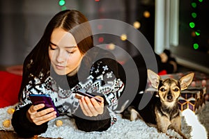 Christmas safe online shopping, a young woman in a winter sweater makes purchases via phone at home online with a small dog, New