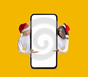 Christmas Sale. Cheerful Young Couple In Santa Hats Pointing At Blank Smartphone