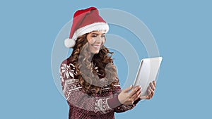 Christmas sale. Pretty lady in woolen sweater and Santa hat shopping online on tablet computer, blue background