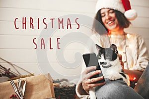 Christmas Sale text sign on cat looking at phone screen with funny emotions and sitting on happy girl legs in christmas lights.
