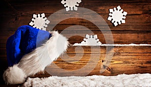 Christmas Santa Hat hanging on Wooden Board Sign. Blue Claus Cap over brown Grunge Wood Background with Snow and Snowflakes. Xmas