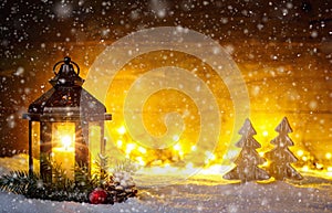 Christmas scene with a lantern, trees, fir branch, snow flakes and blurred lights in front of an illuminated dark wooden board as