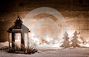 Christmas scene with a lantern, trees, fir branch, snow flakes and blurred lights in front of an illuminated dark wooden board as