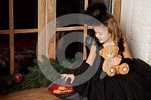 Christmas scene. portrait of a little blond girl, in a black angel costume stealing Santa`s cookies near the window.