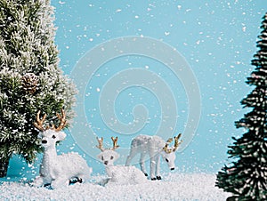 Christmas scene with white reindeer toys and pine trees covered with snow against vivid blue background. Snowy weather