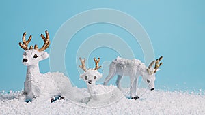 Christmas scene with white reindeer toys and pine trees covered with snow against vivid blue background. Snowy weather