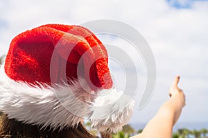 Christmas sea holiday. Back view of happy woman in santa claus hat showing thumb up and relaxing on paradise beach island