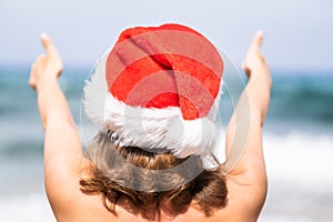 Christmas sea holiday. Back view of happy woman in santa hat showing thumb up and relaxing on paradise beach island getaway.