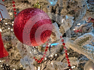 Christmas season 2019. Red shiny decorative ball and red pearls on a Christmas tree covered in snow and cozy yellow lights in the