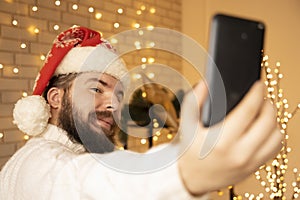 Christmas selfie by middle adult bearded man with white sweater and red hat in warm and cozy decorated room with yellow garland
