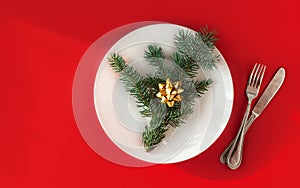 Christmas served dinner, meal table design on red background. White plate, golden bow, fir, fork, knife. New year minimal concept