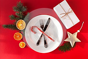 Christmas served dinner, table design on red background. White plate, glass, fork, knife, fir, orange, candy cane and decorations