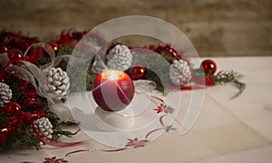 Christmas setting: a red lit candle with cross screen effect on foreground surrounded by pine branches, red baubles, red and white