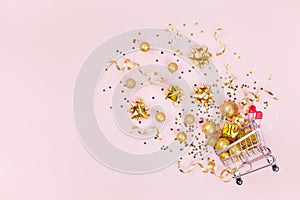 Christmas shopping cart with gift, holiday decorations and golden confetti on pink pastel background top view. Flat lay style.