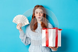 Christmas and shopping concept. Excited redhead girl looking at camera, holding big New Year gift and dollars, buying