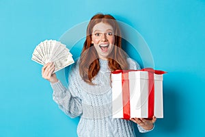 Christmas and shopping concept. Happy redhead woman holding money and big xmas present, showing dollars and gift