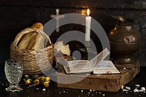 A Christmas table, bread in a basket and a glass of water bring a prayer book to a dark wooden table. Cross in the background