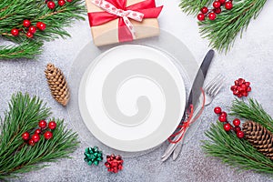 Christmas table place setting with empty white plate, gift box, cutlery with festive decorations on stone background