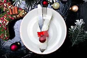 Christmas table place setting. Holidays background. White plate, knife and fork with christmas decorations on black table. Top vie