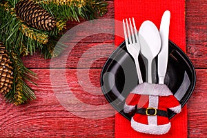 Christmas table place setting with red napkin, black plate, white fork, spoon and knife, decorated Santa jacket and
