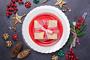 Christmas table place setting with red plate, gift box, cutlery with festive decorations star bow on stone background
