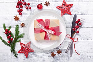 Christmas table place setting with white plate and gift box, cutlery with festive decorations on wooden background