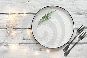 Christmas table setting. Empty plate, garland lights, champagne glass and cutlery. White old wooden table. Top view, flat lay