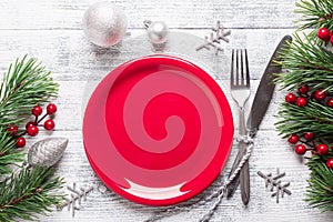 Christmas table setting with empty red plate, gift box and silverware on light wood background. Fir tree branch.