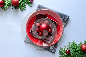 Christmas table setting with red plate, brown napkin in lotus shape and balls on gray wooden background. Holiday decorations