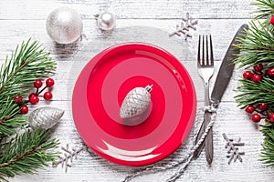 Christmas table setting with red plate and silverware on light wood background. Fir tree branch, holly berries.