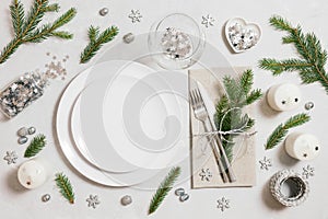 Christmas table setting with silver decorations and fir branches on gray background. Empty white plate for design. Top view, flat