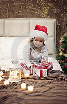 It is Christmas Time! Happy Funny Child Girl Sitting In Bed And Opening The Present On Christmas Morning. Christmas Tree and Light