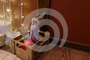 Christmas toy, Santa Claus or festive gnome in hat in wooden box with red bauble for fir-tree. Shiny garland lights, copy space.