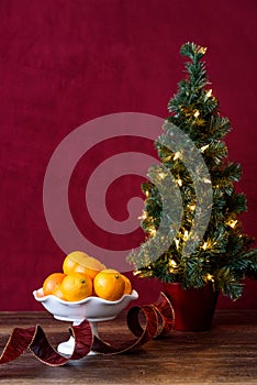 Christmas treat, satsuma oranges in white bowl, with artificial Christmas tree and white lights, red ribbon, wood table, red backg