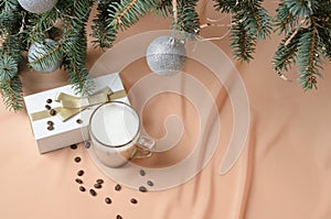 Christmas tree decorated balls, a gift box, a glass mug with coffee and milk on the background of flesh-colored waves